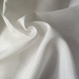 May include: Close-up of white fabric with a subtle textured weave. The fabric is folded and draped, creating soft shadows and highlights. The material appears to be lightweight and possibly linen or cotton.