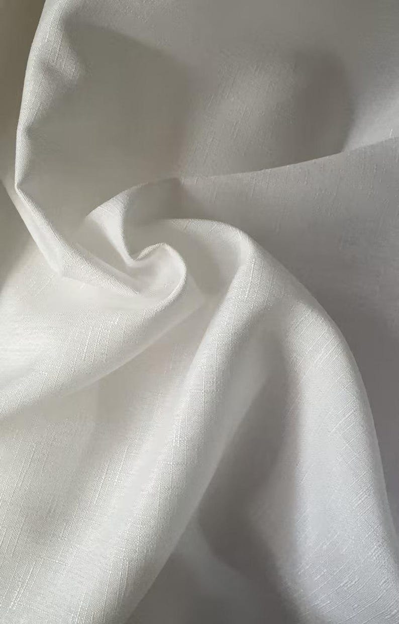 May include: Close-up of white fabric with a subtle textured weave. The fabric is folded and draped, creating soft shadows and highlights. The material appears to be lightweight and possibly linen or cotton.