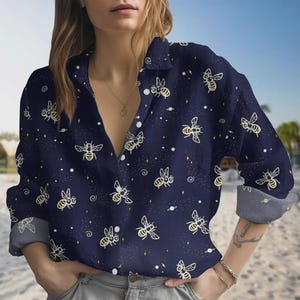 May include: Navy blue button-down shirt featuring a repeating pattern of yellow and white bees, stars, and planets. The long-sleeved shirt has rolled-up cuffs and a collared neckline. The shirt is made of a soft, comfortable material.