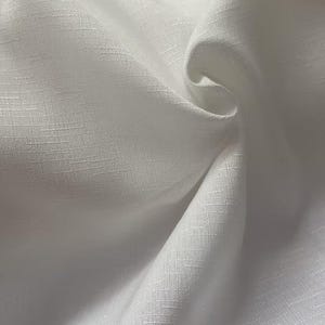 May include: Close-up of white fabric with a textured weave. The fabric is draped, creating soft folds and shadows. The material appears to be lightweight and possibly linen or a similar breathable textile.