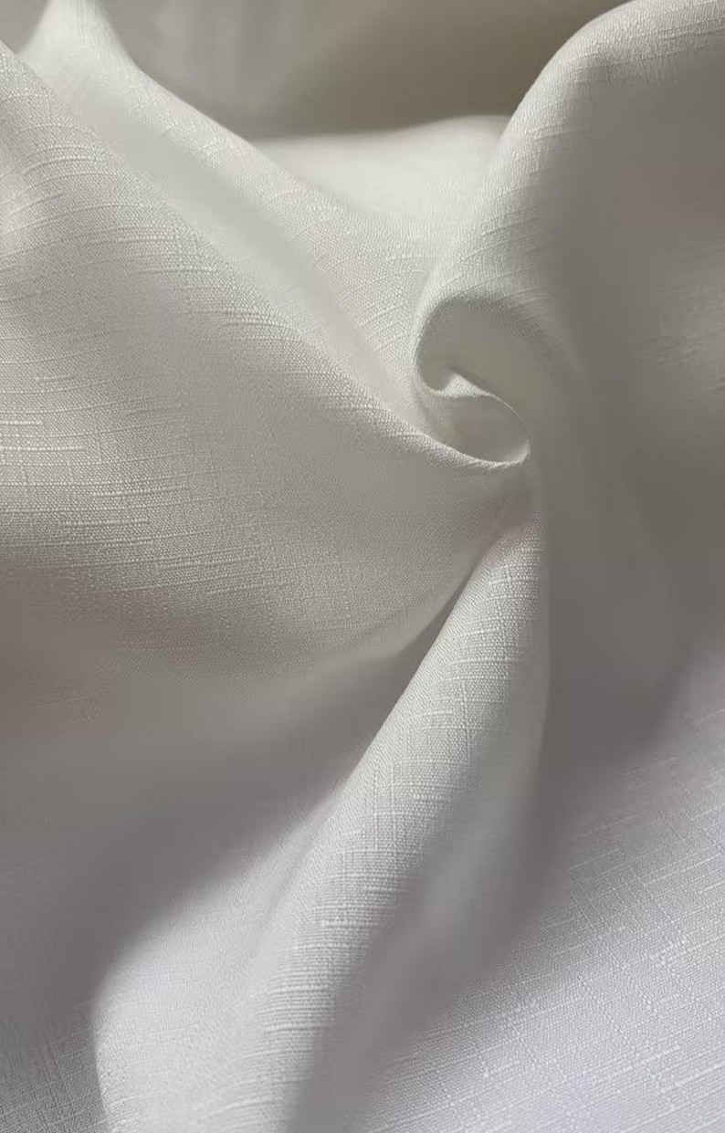 May include: Close-up of white fabric with a textured weave. The fabric is draped, creating soft folds and shadows. The material appears to be lightweight and possibly linen or a similar breathable textile.