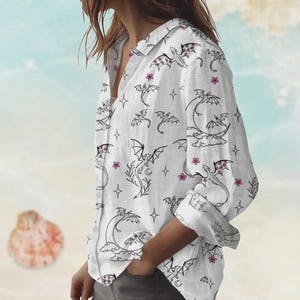 May include: White button-down shirt with a dragon and floral pattern. The long-sleeved shirt features black dragon illustrations, pink flowers, and star accents. The dragon wings have a pink and white pattern.
