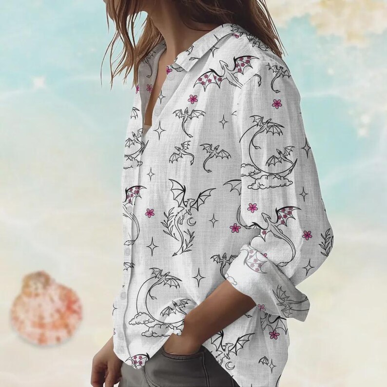 May include: White button-down shirt with a dragon and floral pattern. The long-sleeved shirt features black dragon illustrations, pink flowers, and star accents. The dragon wings have a pink and white pattern.