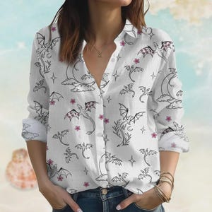 May include: White button-down shirt with a black dragon and pink flower pattern. The long-sleeved shirt features a classic collar and is made of a lightweight fabric. The dragon design includes a crescent moon and stars.