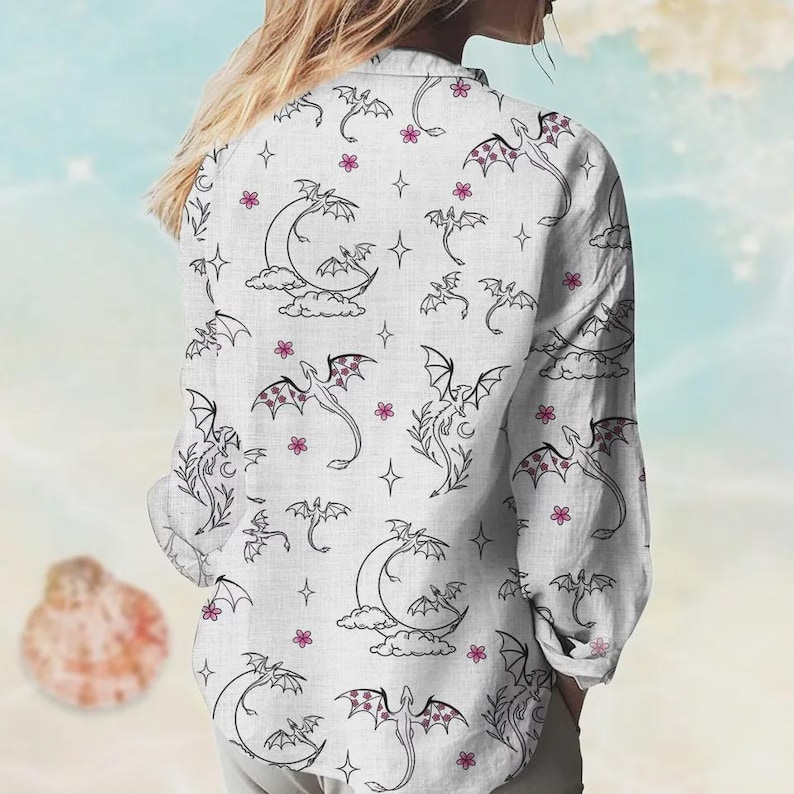 May include: White button-down shirt with a repeating pattern of black dragons, moons, clouds, stars, and pink flowers. The long-sleeved shirt has a classic collar and is made of a lightweight fabric.