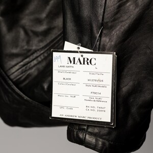 K&ouml;nnte beinhalten: Schwarze Lederjacke mit einem Etikett mit der Aufschrift "MARC". Das Etikett enth&auml;lt auch die W&ouml;rter "LAMB NAPPA" und "BLACK". Die Jacke hat die Gr&ouml;&szlig;e L. Das Etikett enth&auml;lt auch die Modellnummer M1275VA26.