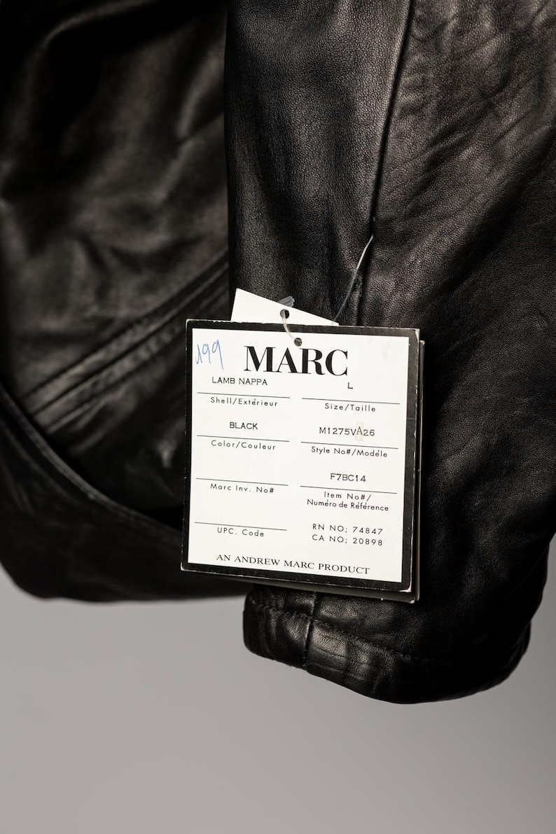 K&ouml;nnte beinhalten: Schwarze Lederjacke mit einem Etikett mit der Aufschrift "MARC". Das Etikett enth&auml;lt auch die W&ouml;rter "LAMB NAPPA" und "BLACK". Die Jacke hat die Gr&ouml;&szlig;e L. Das Etikett enth&auml;lt auch die Modellnummer M1275VA26.