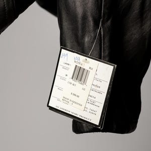 K&ouml;nnte beinhalten: Schwarze Lederjacke mit einem angebrachten Etikett. Das Etikett lautet "LAMB LTR JKT LG" und enth&auml;lt einen Barcode und die Marke "Marc". Die Jacke ist in Gr&ouml;&szlig;e L.