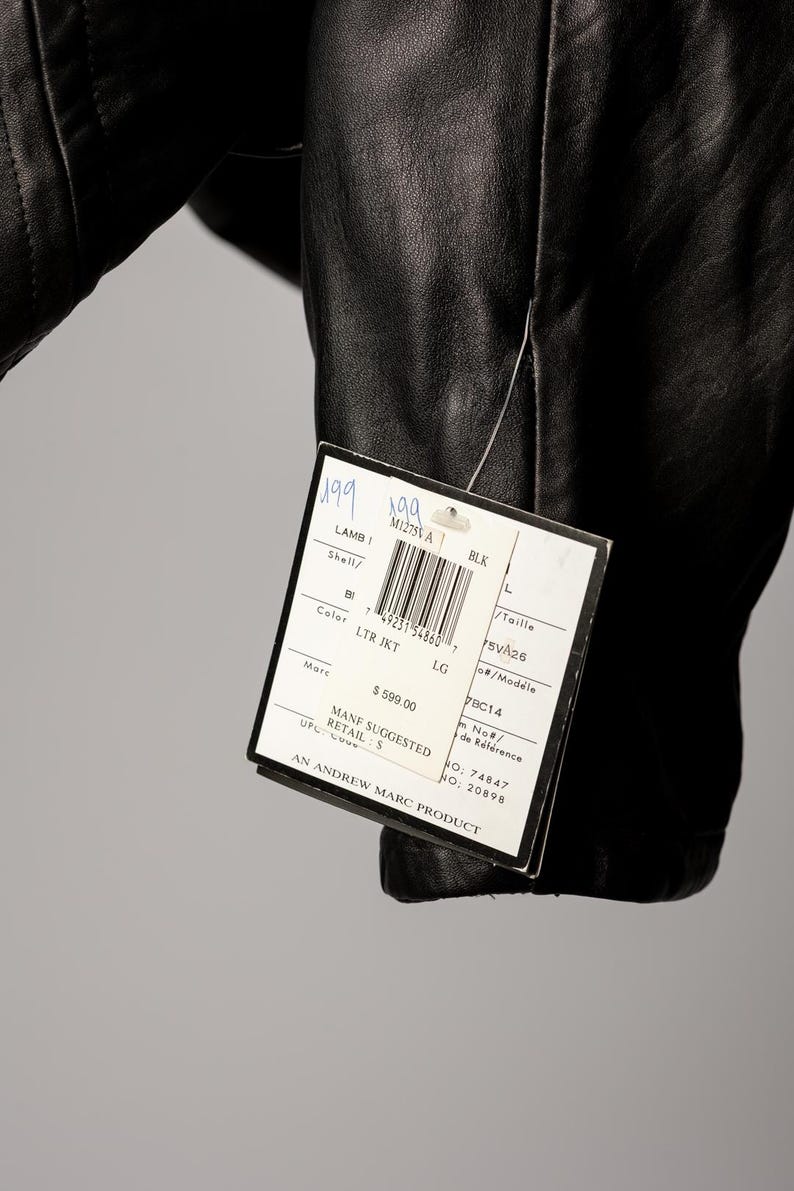 K&ouml;nnte beinhalten: Schwarze Lederjacke mit einem angebrachten Etikett. Das Etikett lautet "LAMB LTR JKT LG" und enth&auml;lt einen Barcode und die Marke "Marc". Die Jacke ist in Gr&ouml;&szlig;e L.