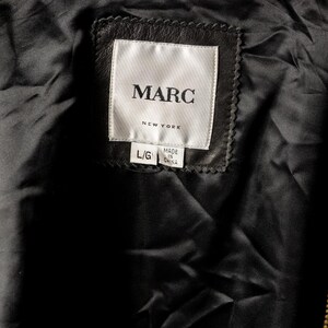 K&ouml;nnte beinhalten: Nahaufnahme des Innenraums einer schwarzen Lederjacke. Das Etikett lautet "MARC NEW YORK" mit den Etiketten "L/G" und "MADE IN CHINA". Das Futter der Jacke ist schwarz und gl&auml;nzend.