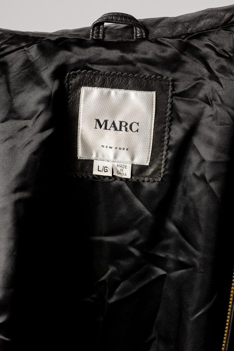 K&ouml;nnte beinhalten: Nahaufnahme des Innenraums einer schwarzen Lederjacke. Das Etikett lautet "MARC NEW YORK" mit den Etiketten "L/G" und "MADE IN CHINA". Das Futter der Jacke ist schwarz und gl&auml;nzend.
