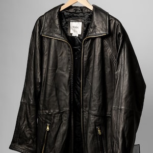 K&ouml;nnte beinhalten: Schwarze Lederjacke mit Rei&szlig;verschluss vorne, zwei Vordertaschen mit Rei&szlig;verschl&uuml;ssen und einem Kragen. Die Jacke h&auml;ngt an einem Holzb&uuml;gel. Das Etikett in der Jacke lautet "MARC".