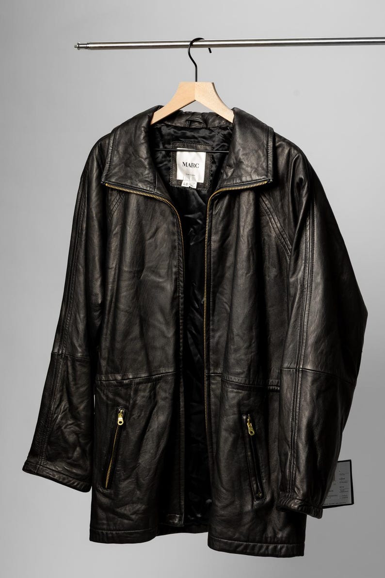 K&ouml;nnte beinhalten: Schwarze Lederjacke mit Rei&szlig;verschluss vorne, zwei Vordertaschen mit Rei&szlig;verschl&uuml;ssen und einem Kragen. Die Jacke h&auml;ngt an einem Holzb&uuml;gel. Das Etikett in der Jacke lautet "MARC".