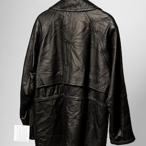 K&ouml;nnte beinhalten: Schwarze Lederjacke an einem Holzkleiderb&uuml;gel. Die Jacke hat ein klassisches Design mit Kragen, Taschen und Knopfleiste. Das Leder scheint leicht abgenutzt zu sein, was ihm einen Vintage-Look verleiht. Ein Etikett ist an der Jacke befestigt.