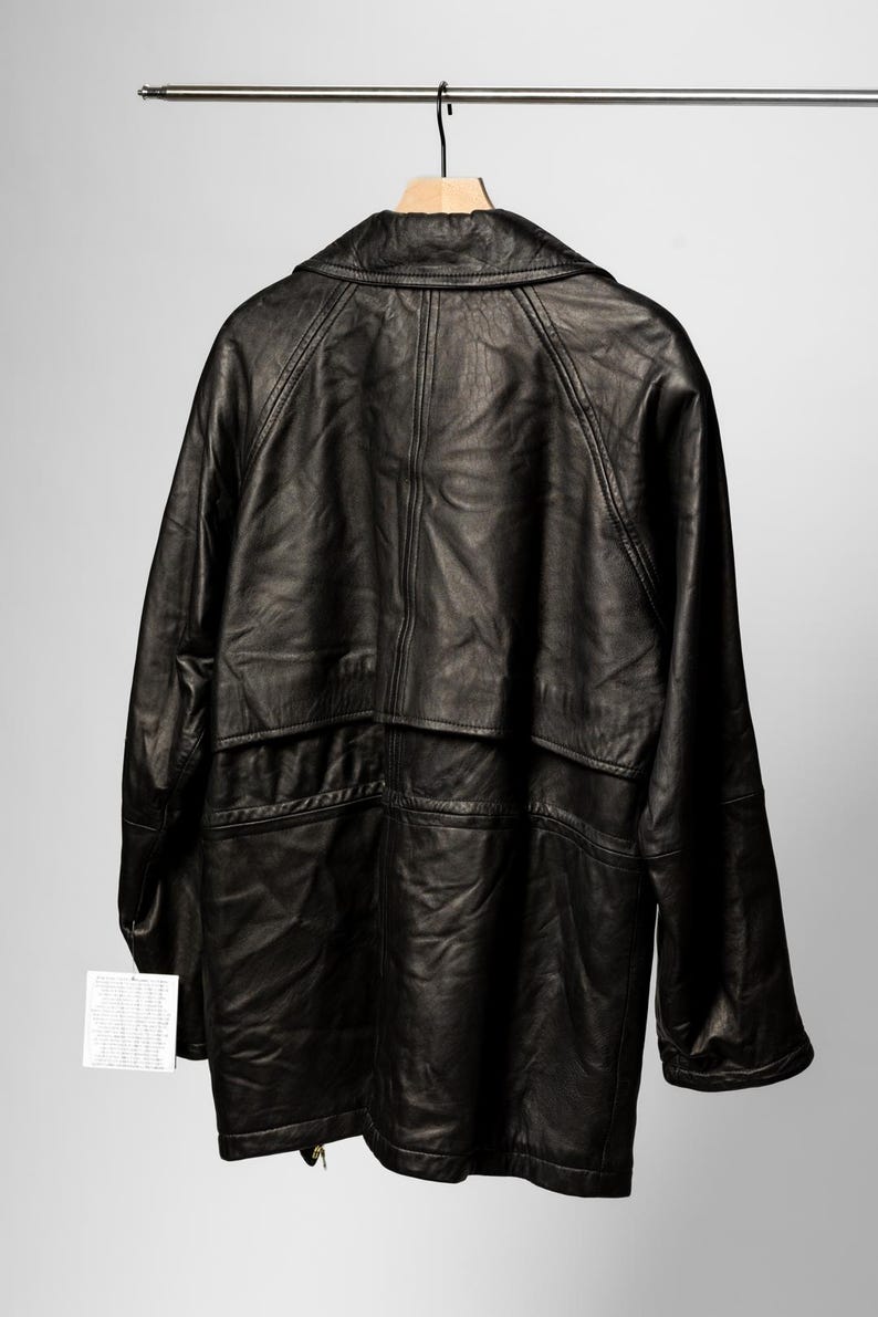 K&ouml;nnte beinhalten: Schwarze Lederjacke an einem Holzkleiderb&uuml;gel. Die Jacke hat ein klassisches Design mit Kragen, Taschen und Knopfleiste. Das Leder scheint leicht abgenutzt zu sein, was ihm einen Vintage-Look verleiht. Ein Etikett ist an der Jacke befestigt.