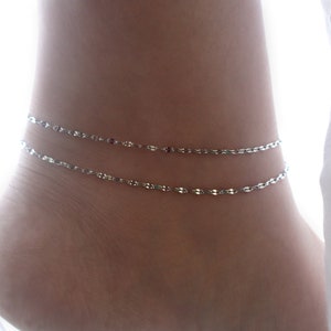 May include: A silver anklet with two delicate chains. The anklet is made of a thin, linked chain with small, round beads.