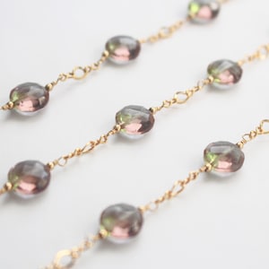 May include: Close-up of three strands of delicate jewellery. Each strand features faceted, oval-shaped beads in shades of pink, green, and grey, strung on a gold-coloured chain. The chains are arranged diagonally across a white background.