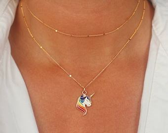Könnte beinhalten: Eine goldene Kettenkette mit einem Regenbogen-Einhorn-Anhänger. Das Einhorn ist weiß mit einem goldenen Horn und einer Regenbogenmähne und einem Regenbogen-Schwanz.