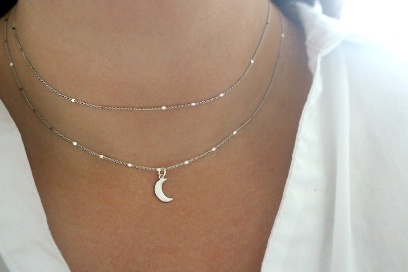May include: A silver chain necklace with a crescent moon pendant. The necklace is layered with a second, shorter chain.