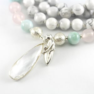 May include: A multi-strand necklace with white, pink, and green beads, and a silver pendant featuring a pair of figures holding a teardrop-shaped crystal.