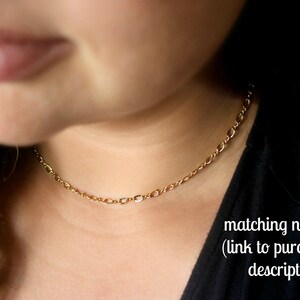 May include: A close-up of a woman wearing a gold chain necklace. The necklace is a delicate, simple chain with a subtle shine. The woman is wearing a black shirt and has long brown hair.