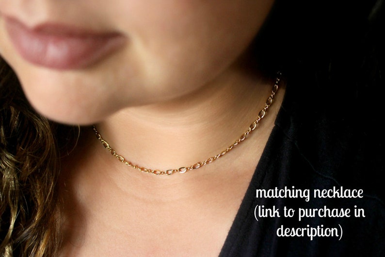 May include: A close-up of a woman wearing a gold chain necklace. The necklace is a delicate, simple chain with a subtle shine. The woman is wearing a black shirt and has long brown hair.