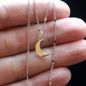 May include: A silver chain necklace with a crescent moon pendant. The chain is delicate and features small, round links. The pendant is simple and elegant.