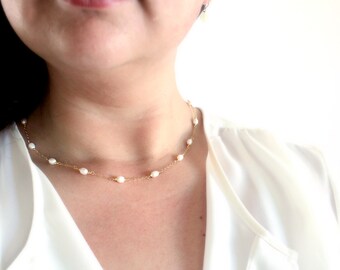 Op de afbeelding: Een delicate gouden ketting met kleine witte parels. De ketting wordt gedragen door een persoon met een wit shirt.