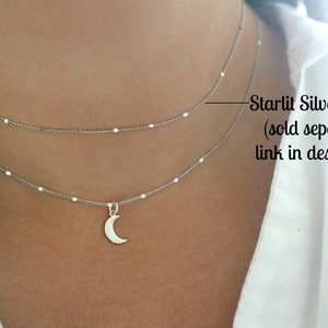 May include: A silver chain necklace with a crescent moon pendant. The necklace is layered with a second, shorter chain. The text "Starlit Silver Choker (sold separately, link in description)" is visible in the image.