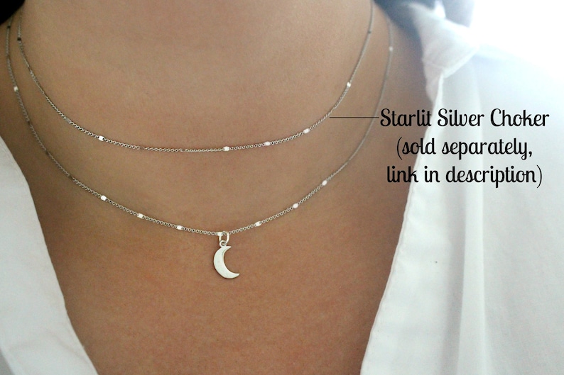 May include: A silver chain necklace with a crescent moon pendant. The necklace is layered with a second, shorter chain. The text "Starlit Silver Choker (sold separately, link in description)" is visible in the image.