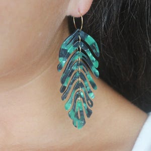 Peut inclure: Gros plan d'une boucle d'oreille en forme de feuille. La boucle d'oreille est dotée d'un cerceau doré et d'un design superposé dans des tons de vert d'eau et de noir. La boucle d'oreille est portée à l'oreille.