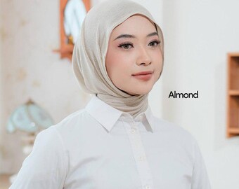 May include: A beige hijab, labeled "Almond", is worn with a white collared shirt. The hijab is draped over the head and neck, creating a soft, flowing appearance. The shirt is buttoned up, with the collar neatly in place.