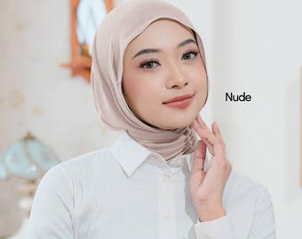 May include: A woman wearing a nude-colored hijab and a white button-down shirt. The word "Nude" is printed in black on the right side of the image. The woman is touching her face with her right hand.