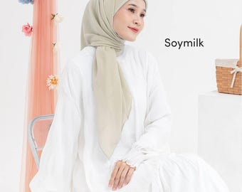 May include: A woman wearing a light beige hijab and a white dress. The hijab is draped over the head and shoulders. The text "Soymilk" is visible on the right side of the image. The background includes a sheer peach-colored curtain with floral accents.