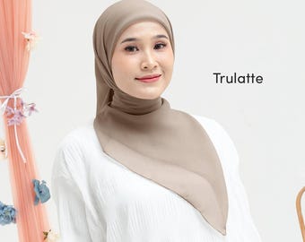 May include: A woman wearing a taupe-colored hijab and a white blouse. The hijab is draped elegantly, covering the head and neck. The word "Trulatte" is printed in black on the right side of the image. The background is a soft, neutral color.