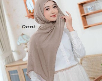 May include: A woman wearing a chestnut-colored hijab. The hijab is draped over the head and shoulders, with the word "Chesnut" printed on the image. She is wearing a white button-down shirt and a white skirt.