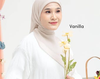 May include: A solid light beige hijab worn over a white long-sleeved top. The word Vanilla is printed in black text to the right of the head. A stem with pale yellow flowers is held in front of the white shirt, with a woven basket visible.