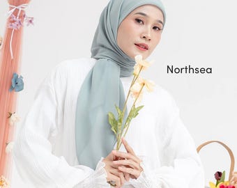 May include: A woman wearing a light blue hijab and a white long-sleeved shirt. The text "Northsea" is printed in black on the right side of the image. She is holding yellow flowers.