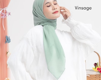 May include: A light green hijab, draped elegantly, with the word "Vinsage" in the upper right corner. The hijab is paired with a white long-sleeved blouse. The background is a soft, neutral color.