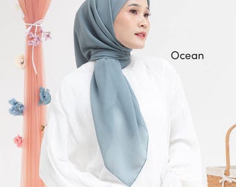 May include: A light blue hijab, draped over the head and shoulders. The hijab is paired with a white blouse. The word "Ocean" is printed in black on the right side of the image. The background includes a decorative pink curtain with floral accents.