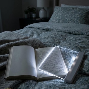 May include: An open book lit by a white LED reading light. The light is rectangular with a handle and shines brightly on the pages. The book is on a bed with a patterned duvet cover and a grey blanket.