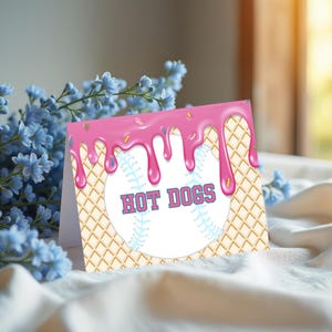 May include: A folded greeting card with a baseball design. The card features a baseball with the words "HOT DOGS" in pink. The top of the card has pink icing dripping down, with sprinkles. The background has a waffle cone pattern.