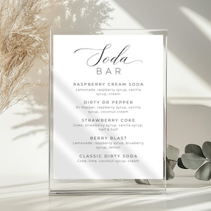 May include: A clear acrylic sign with the words "Soda BAR" in elegant script. Below, a list of soda concoctions with ingredients like raspberry syrup, vanilla syrup, and coconut cream. The sign is displayed in a bright setting.