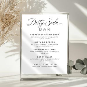 May include: A clear acrylic sign displaying "Dirty Soda BAR" in cursive. The sign details soda recipes such as Raspberry Cream Soda, Dirty Dr Pepper, Strawberry Coke, Berry Blast, and Classic Dirty Soda, with ingredients listed.