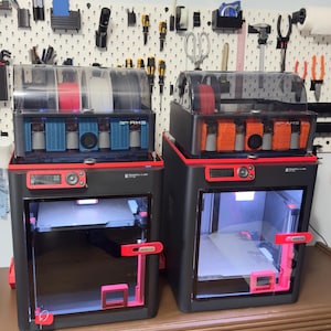 May include: Two black and red 3D printers with clear tops, showcasing spools of filament in various colors. The printers have red accents and are set on a brown surface. A pegboard with tools hangs in the background.