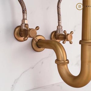 May include: Antique brass-colored plumbing fixtures, including a curved pipe, valves, and connecting hoses, are mounted on a white marble-patterned surface. The fixtures have a vintage aesthetic, with cross-shaped handles on the valves.