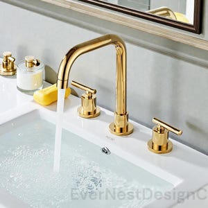 Peut inclure: Un robinet de salle de bain doré avec un bec en arc de cercle et deux poignées à levier. Le robinet est monté sur un évier blanc, avec de l'eau qui coule du bec. Des accessoires dorés supplémentaires sont visibles.