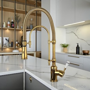 May include: A gold-toned kitchen faucet with a flexible, coiled neck and a separate arched spout. The faucet has a classic design with a white-handled lever. The faucet is installed on a white countertop.