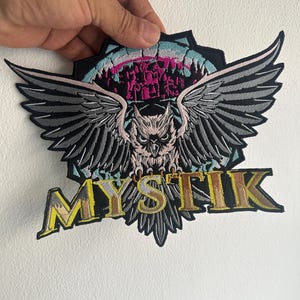 Può includere: Toppa ricamata con un gufo con le ali spiegate, nei toni del grigio, nero e rosa. La parola "MYSTIK" è visualizzata in lettere dorate con un contorno giallo. La toppa ha un bordo nero e una forma unica.