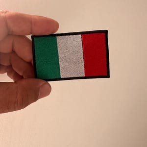 Può includere: Toppa ricamata raffigurante la bandiera italiana. La toppa rettangolare presenta strisce verticali verdi, bianche e rosse, bordate di nero. La toppa è tenuta da una mano su uno sfondo neutro.